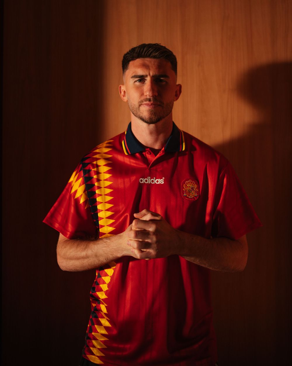 Man wearing red Adidas Spain retro football shirt with geometric pattern, posing in dramatic lighting.