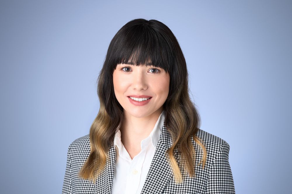 Professional headshot of a smiling young Asian woman with bangs wearing a houndstooth blazer against a blue background.