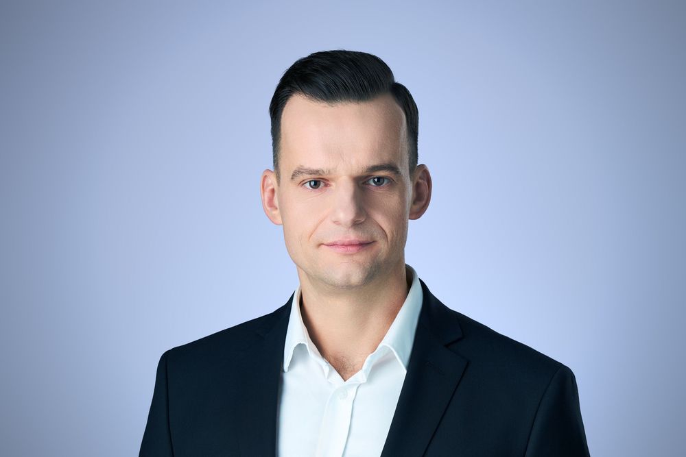 Professional headshot of a young businessman in a dark blazer and white shirt against a light blue background.