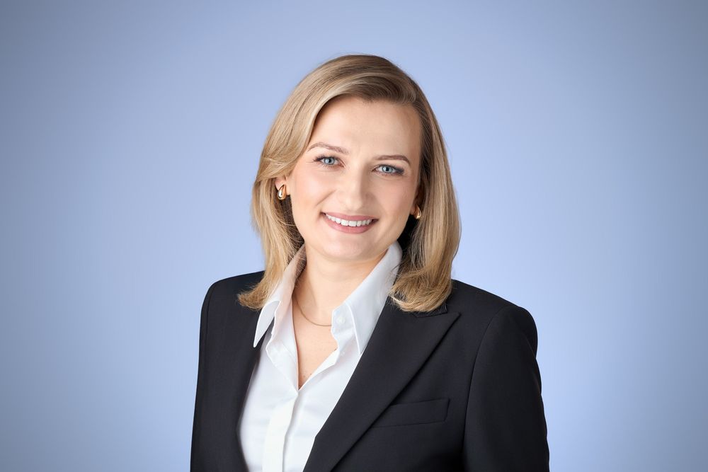 Professional businesswoman in black blazer smiling confidently against a light blue background.