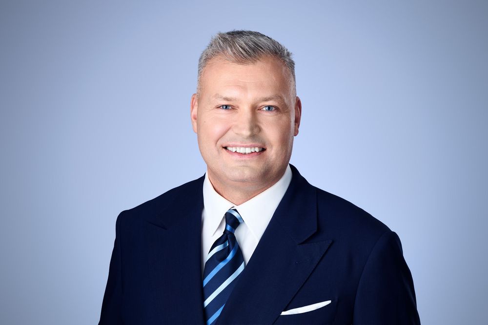 Professional headshot of a smiling man in a navy suit and striped tie against a light blue background.