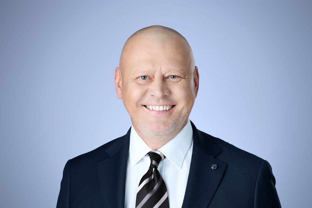 Professional bald man in dark suit and striped tie smiling confidently against light blue background.