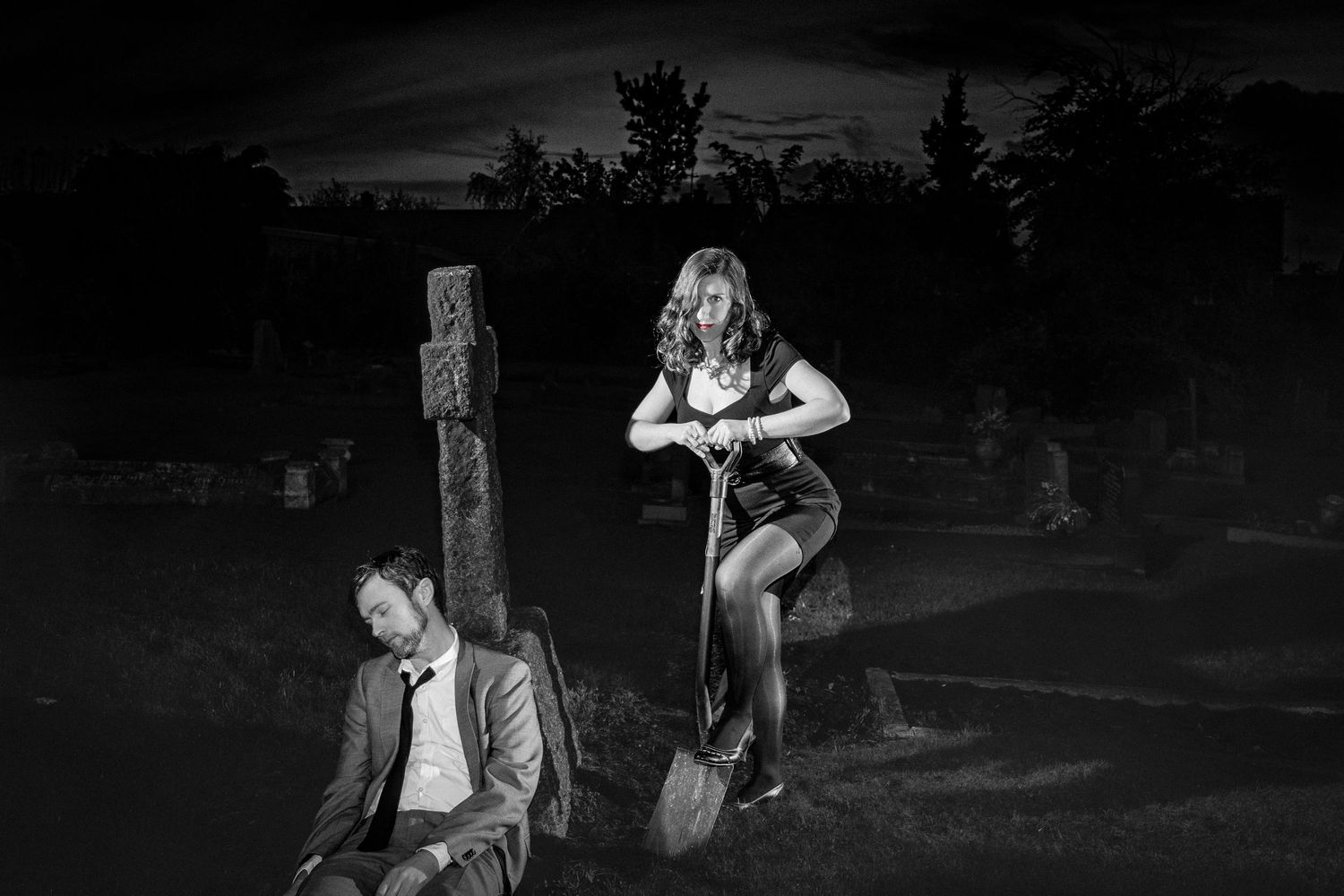Black and white photo of a woman with a shovel standing over a man leaning against a gravestone in a cemetery at night.