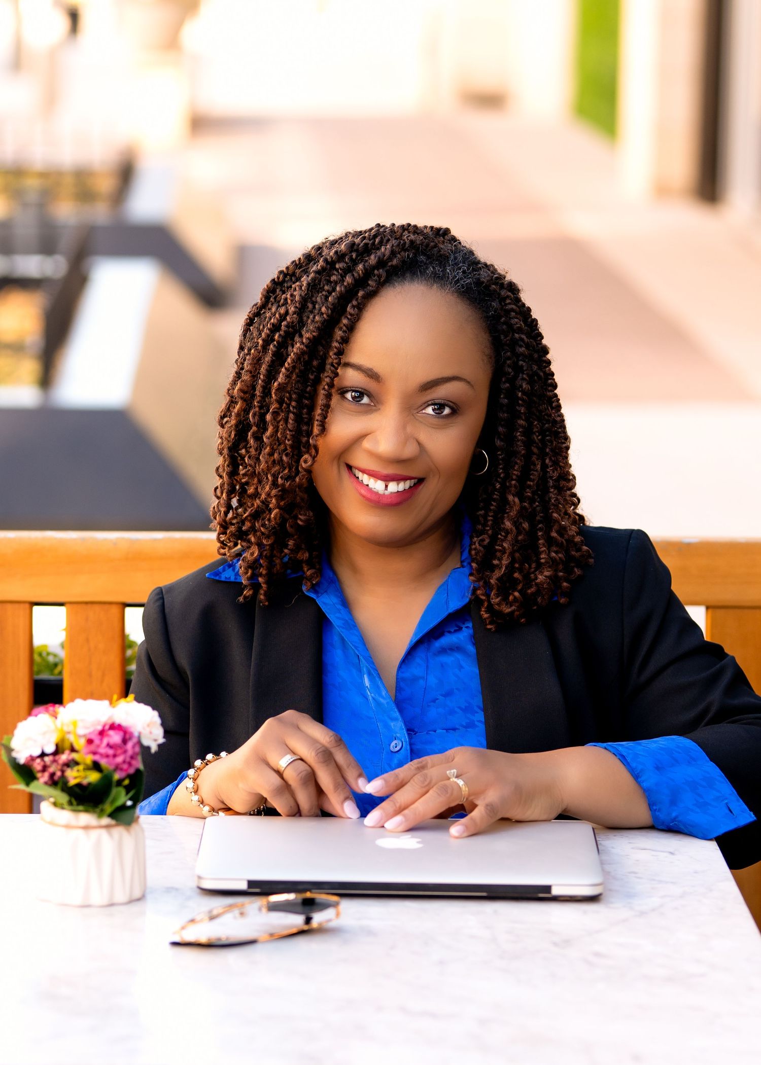 Professional Black woman in blue blouse and blazer smiling at outdoor table with laptop, exuding confidence.