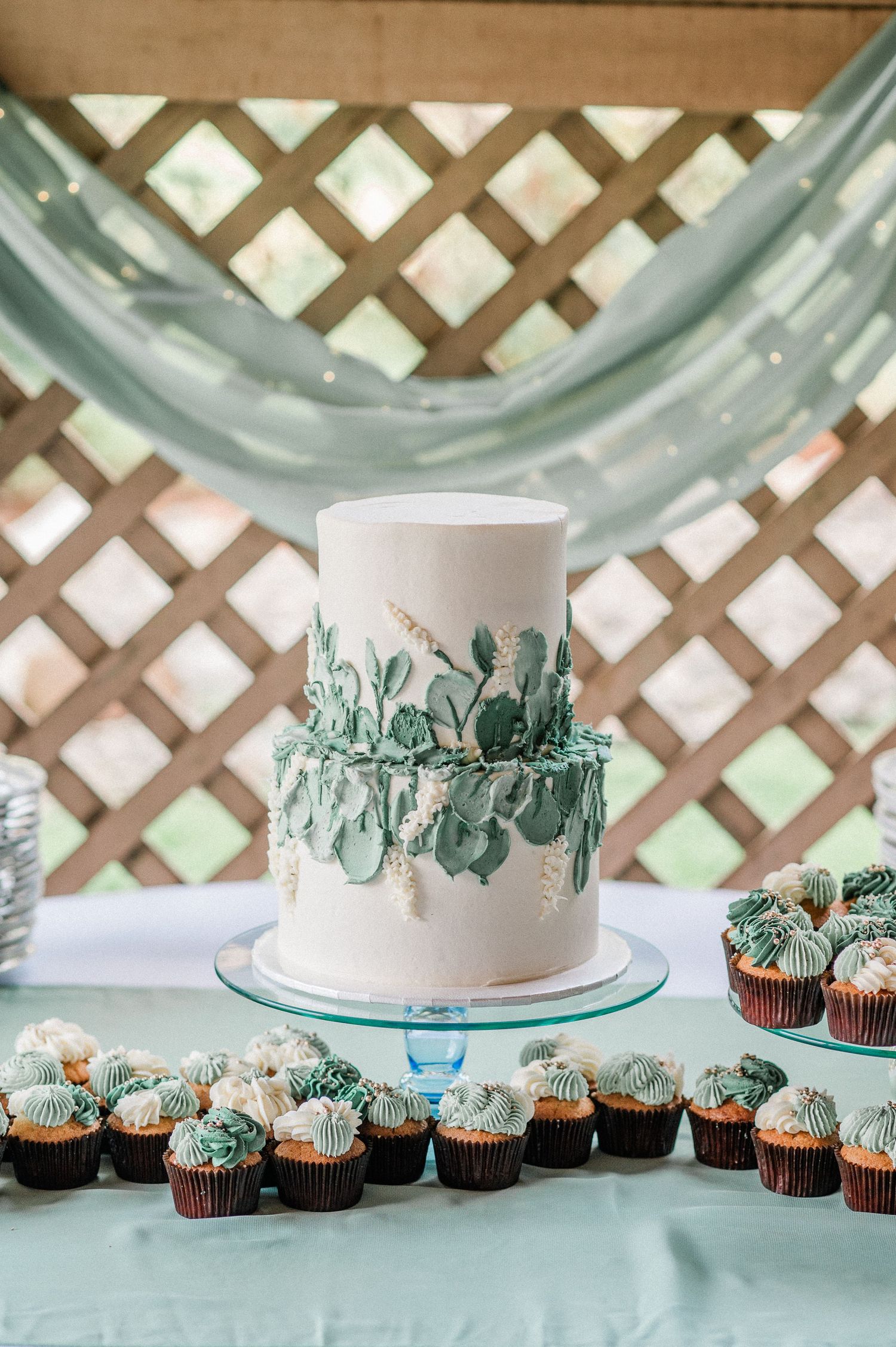 Two-tier white wedding cake with green succulent decorations on a glass stand, surrounded by cupcakes on a teal tablecloth.