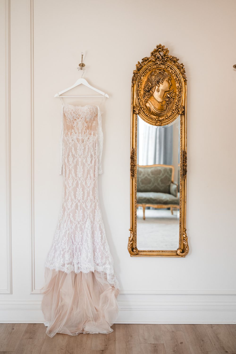Elegant lace mermaid wedding dress hanging beside an ornate gold mirror with cameo detail in a bright white room.