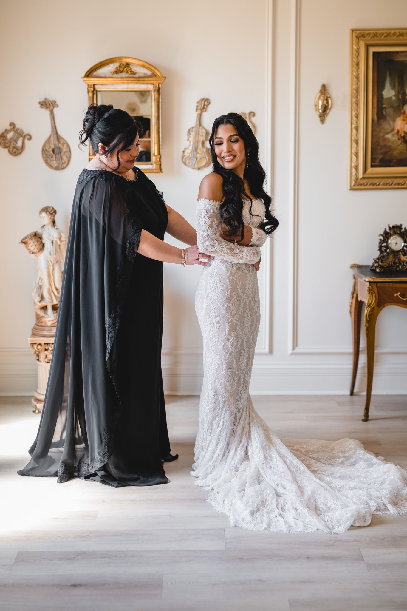 Bride in lace wedding gown being helped by woman in black dress in elegant room with gold decor.