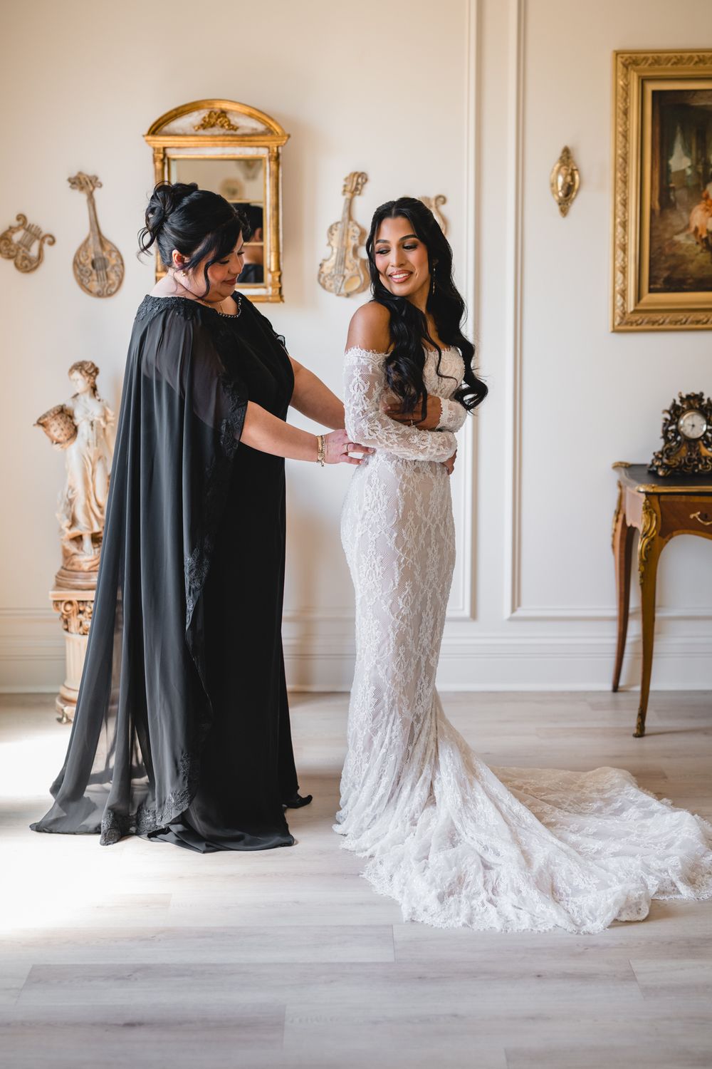 Bride in lace wedding gown being helped by woman in black dress in elegant room with gold decor.