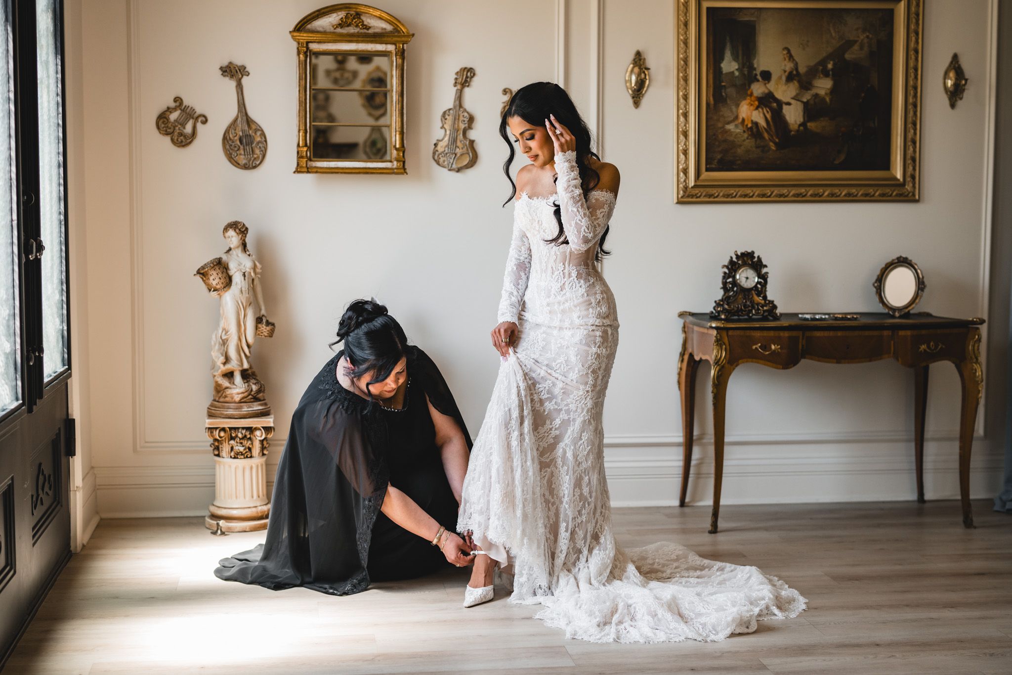 Bride in lace gown gets shoes fastened by assistant in elegant room with antique decor and classical paintings.