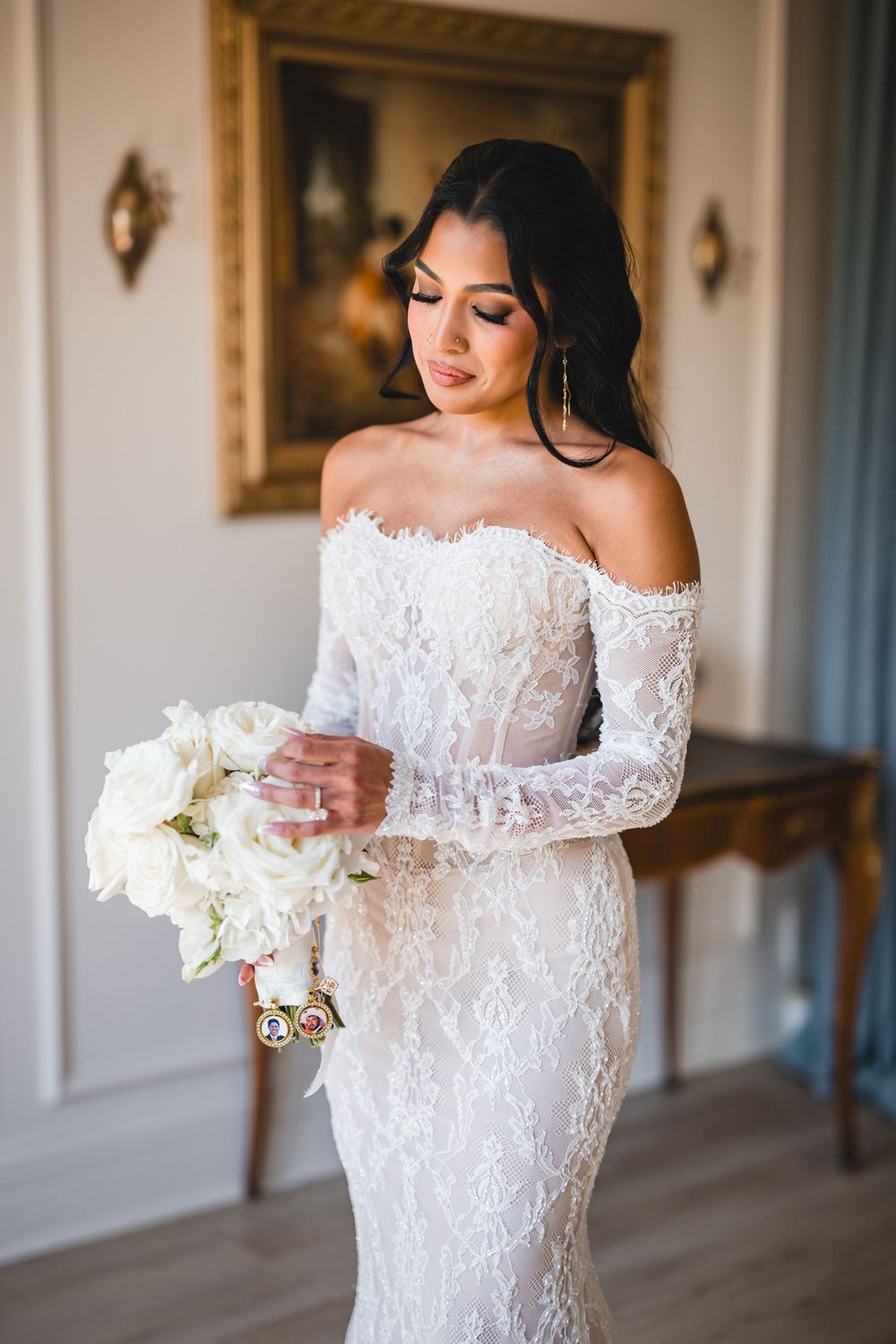 Bride in off-shoulder lace wedding gown holding white rose bouquet in elegant room.