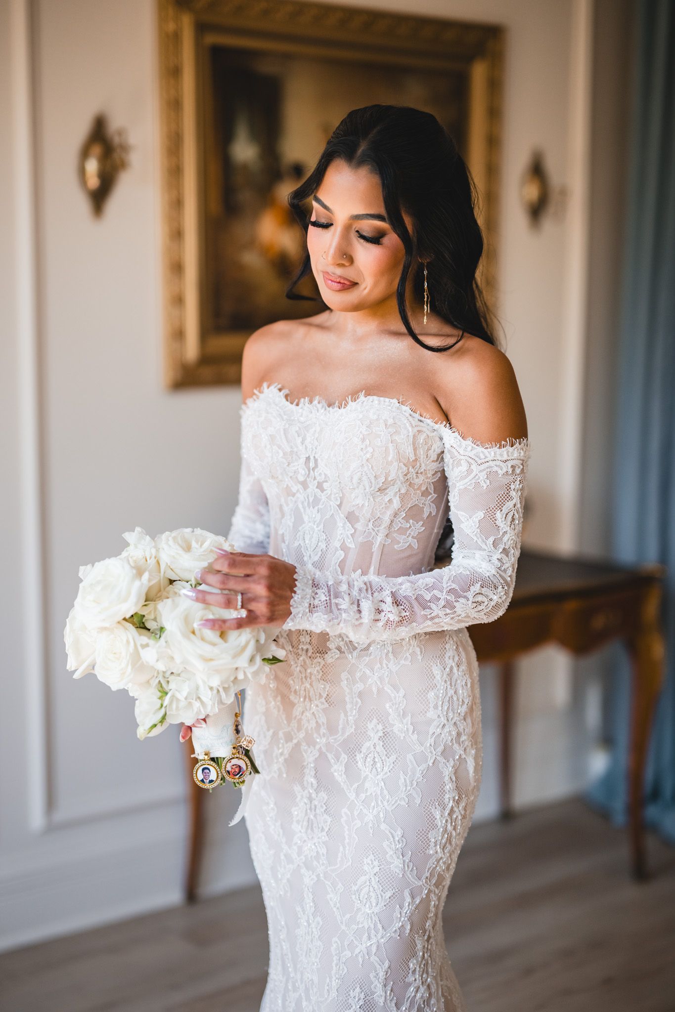 Bride in off-shoulder lace wedding gown holding white rose bouquet in elegant room.
