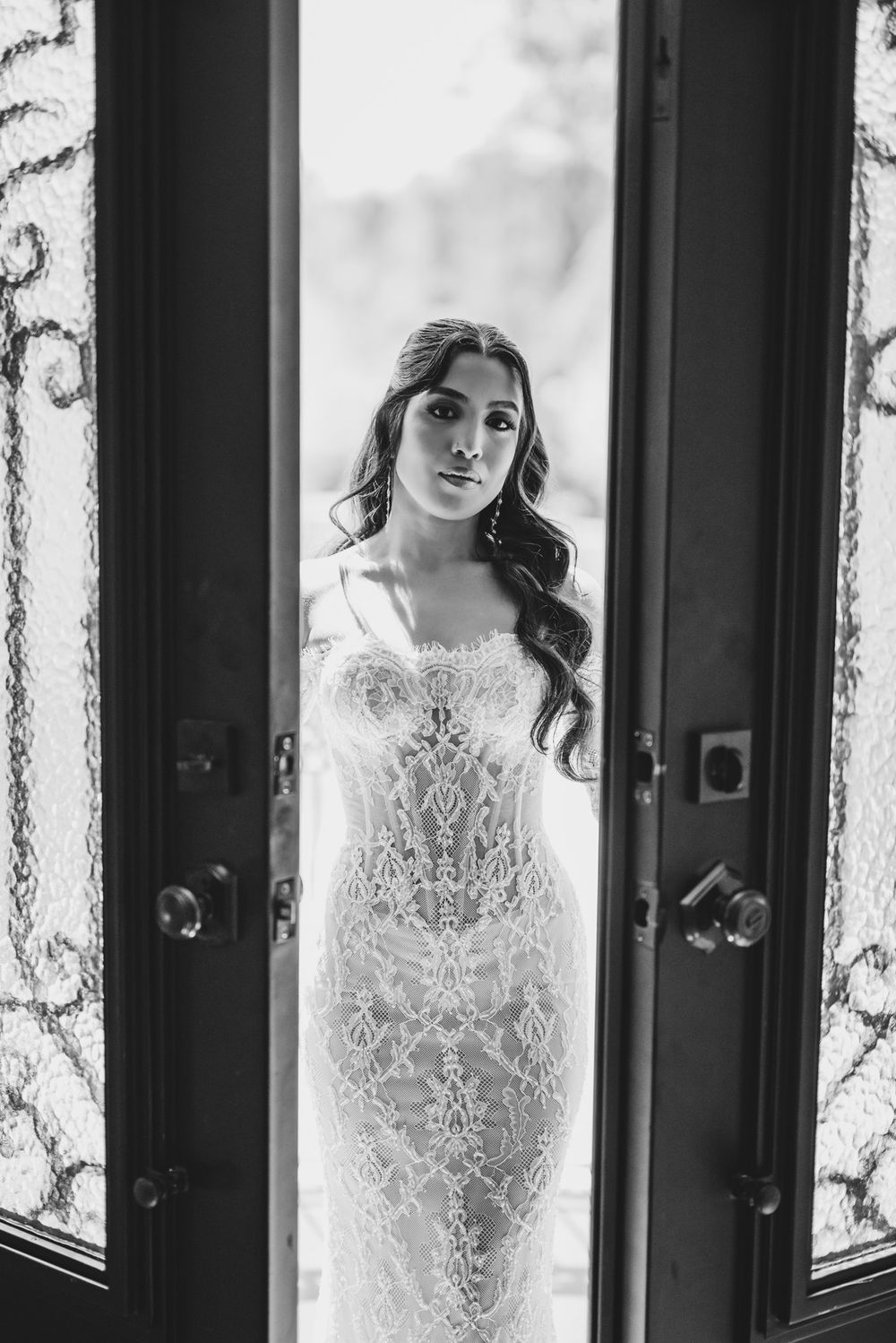 Elegant bride in lace wedding gown standing between ornate glass doors in black and white portrait.