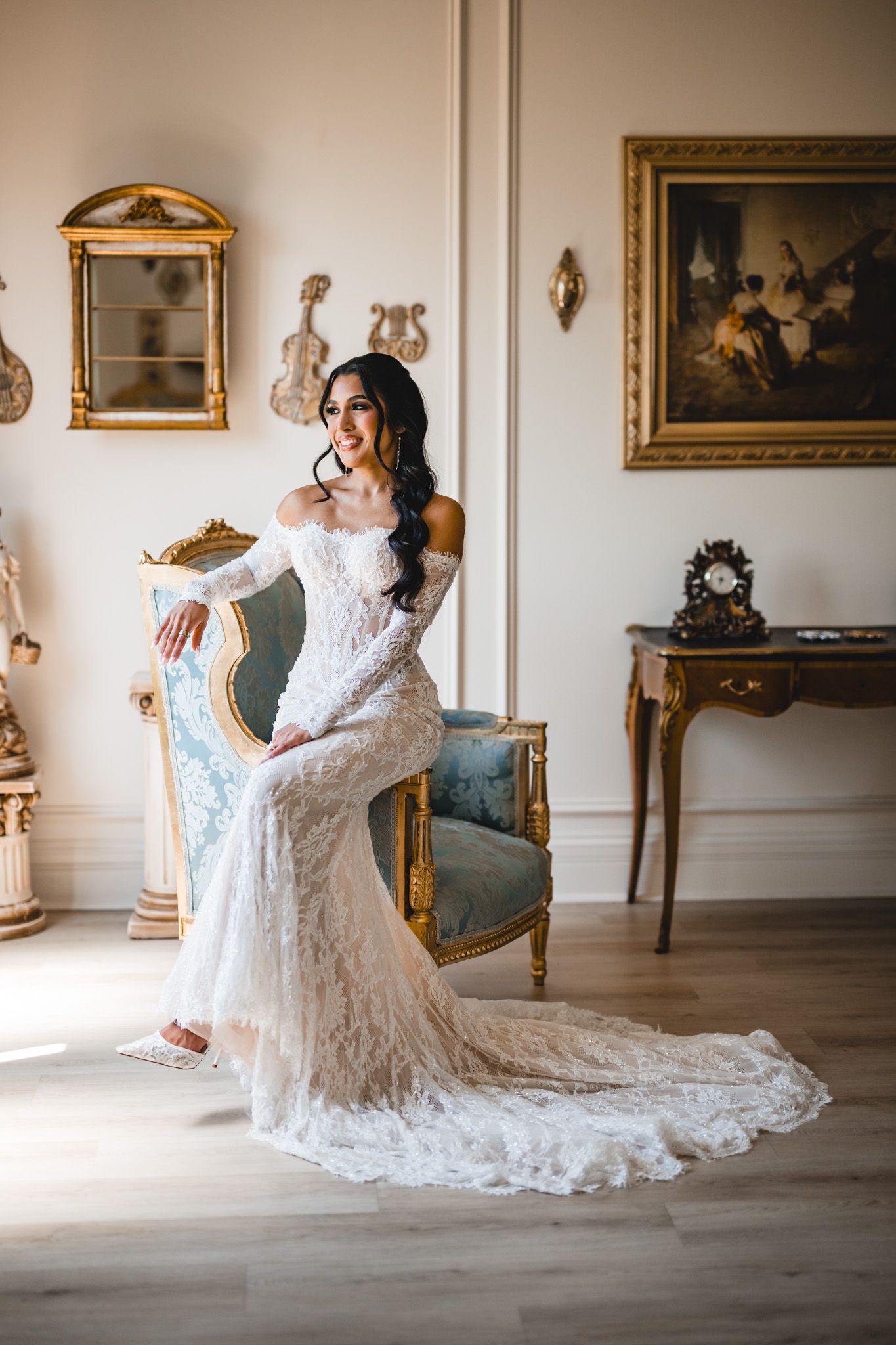 Bride in off-shoulder lace wedding gown with train poses beside ornate gold chair in elegant room.