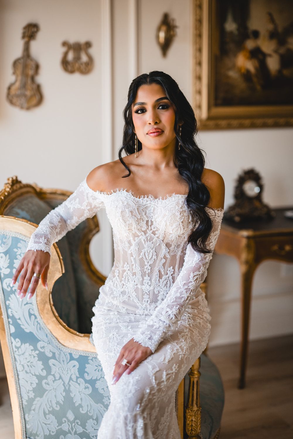Bride in off-shoulder long sleeve lace wedding gown posing beside ornate gold French chair in elegant room.