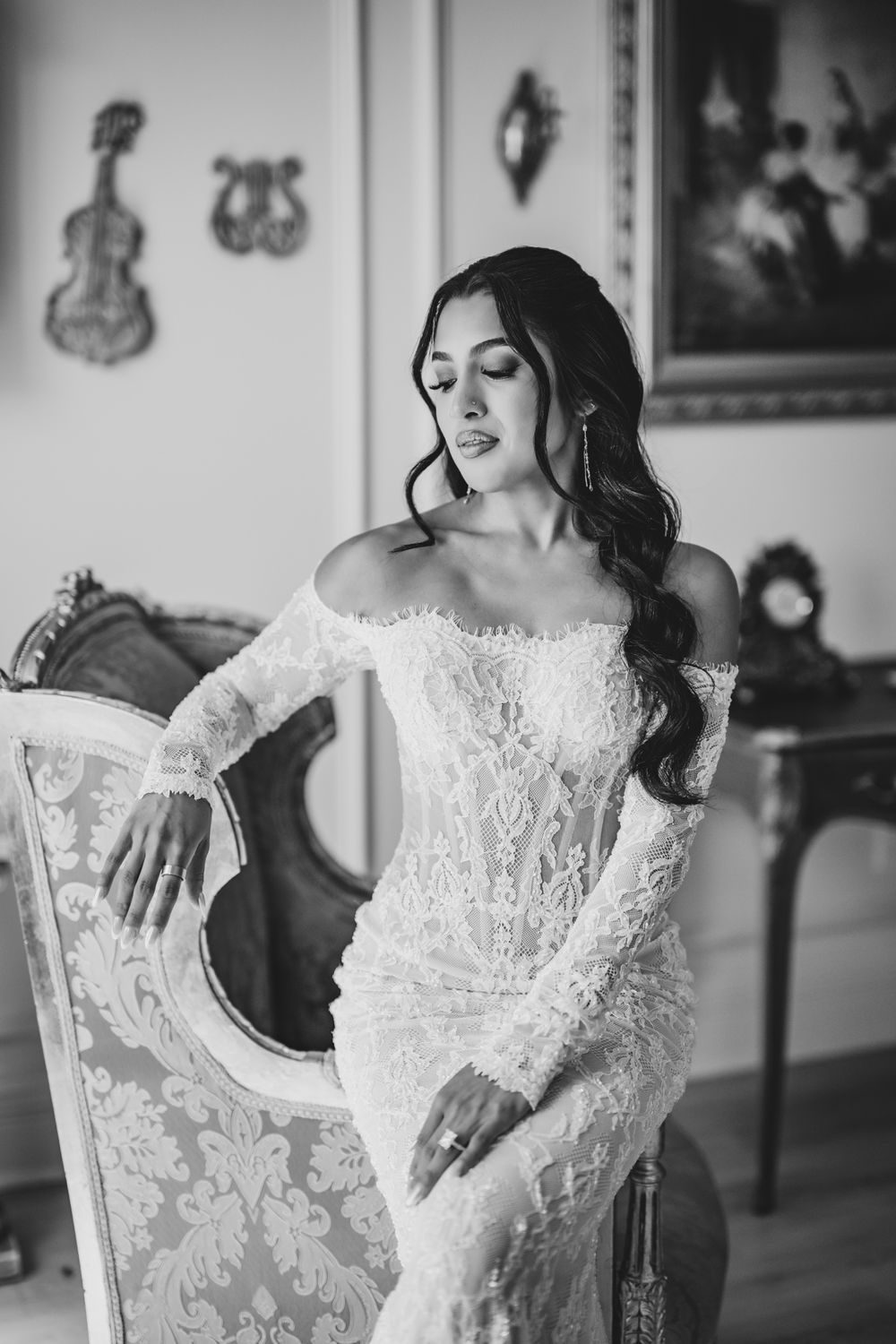 Elegant bride in off-shoulder lace wedding gown poses gracefully in a vintage-styled room, black and white photo.