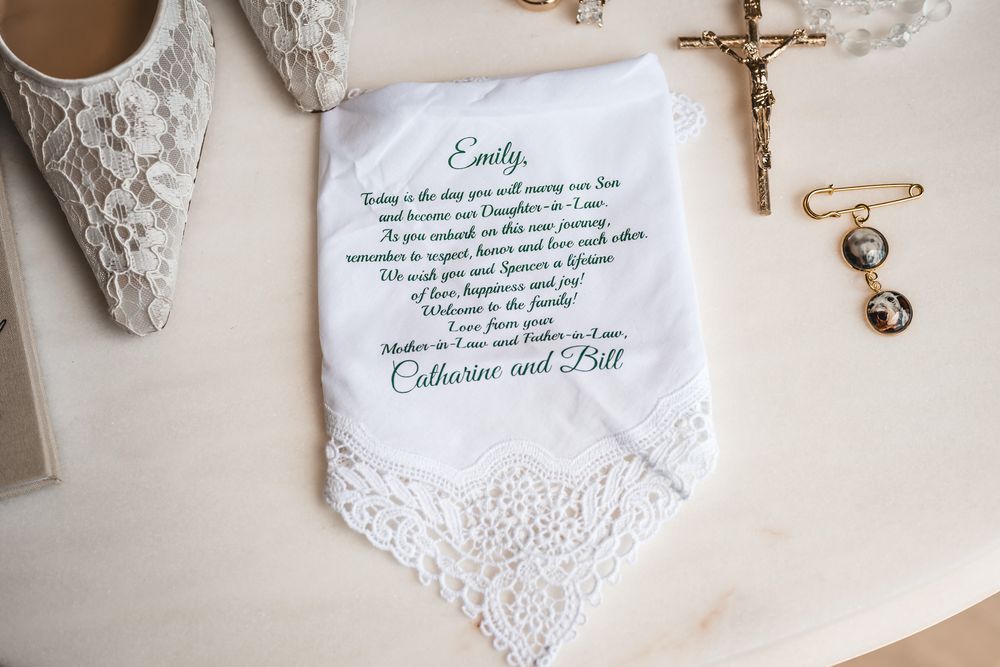 Personalized wedding handkerchief with green script message from parents, displayed beside bridal shoes and gold rosary.