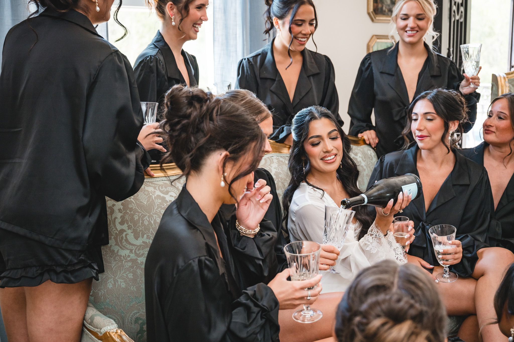 Bridesmaids in black satin robes celebrate with champagne around a smiling bride during wedding morning preparations.