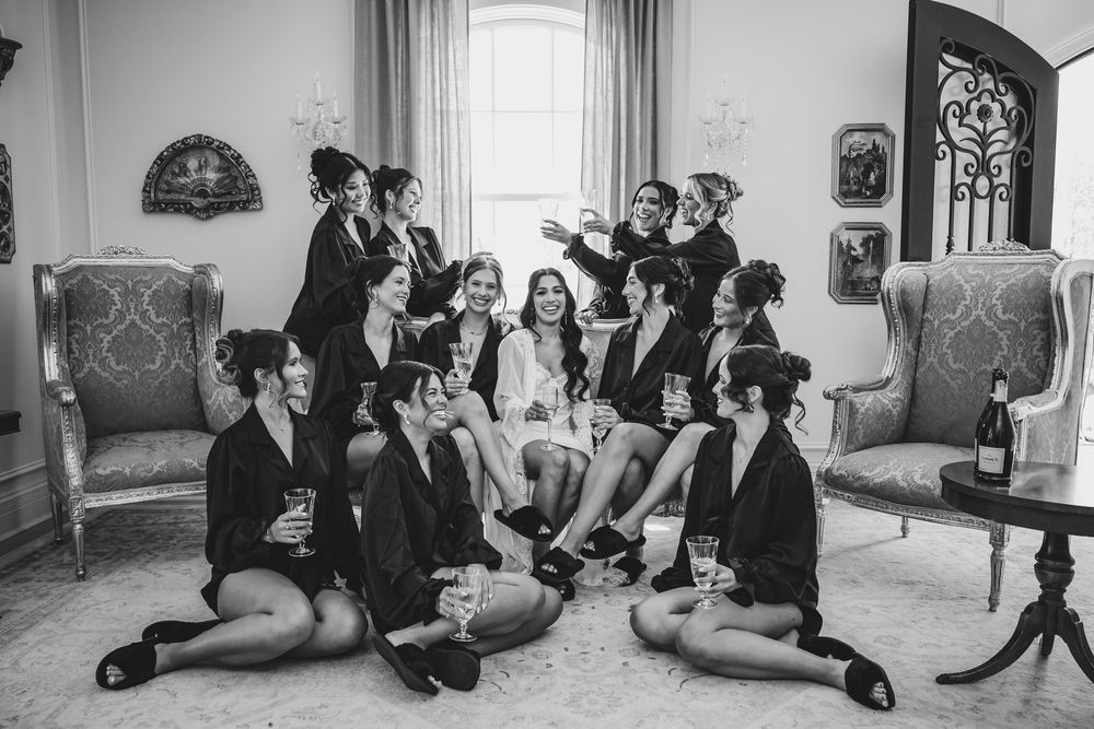 Black and white photo of bride and bridesmaids in robes celebrating with champagne in an elegant room.
