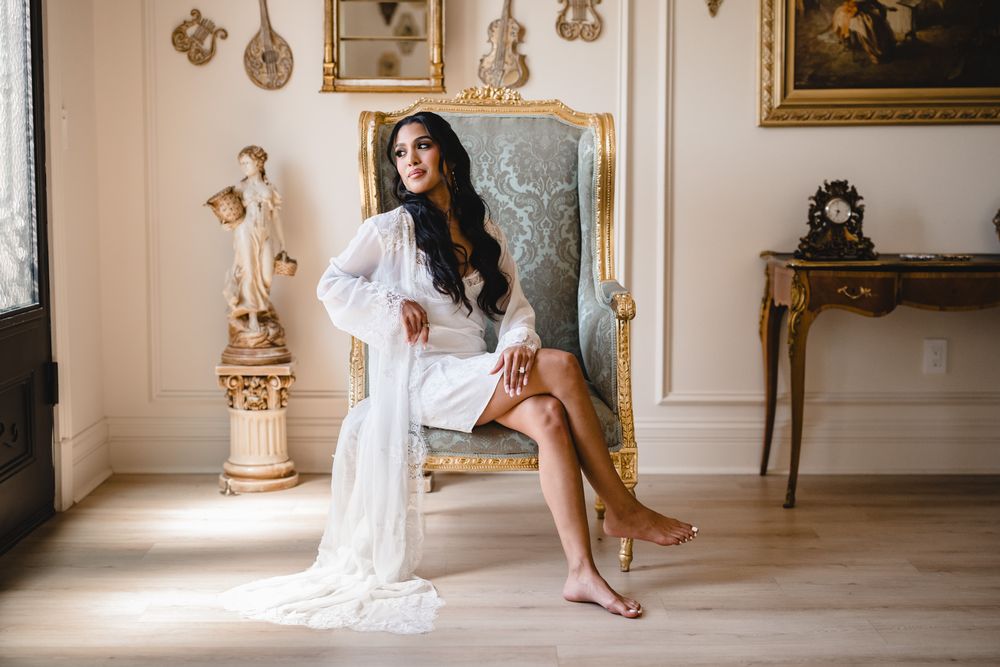 A bride in a white lace robe sits elegantly on a gold ornate chair in a luxurious room with classical decor.