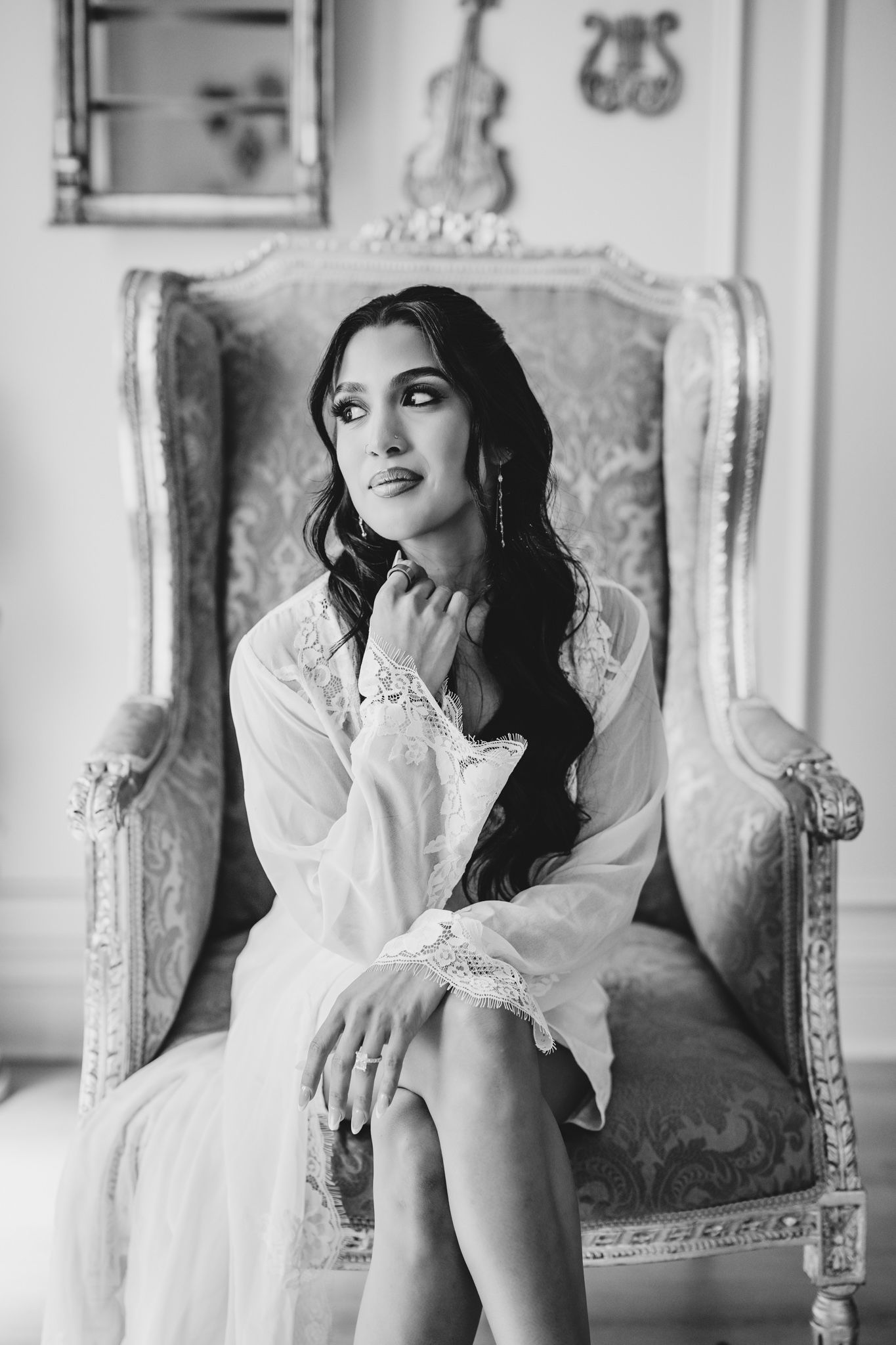 Bride in white lace robe sitting elegantly in ornate vintage armchair, black and white portrait photography.