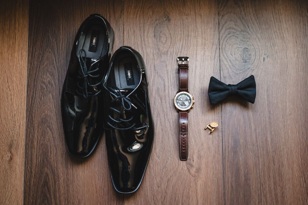 Groom wedding accessories flat lay featuring black patent leather shoes, bow tie, watch, and cufflinks.