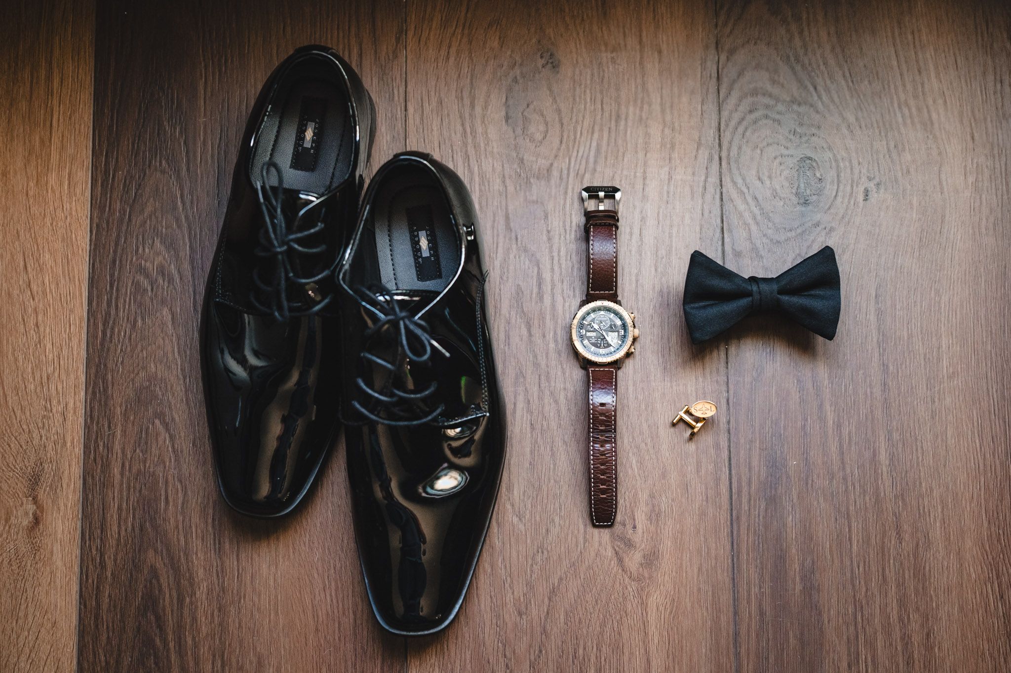 Groom wedding accessories flat lay featuring black patent leather shoes, bow tie, watch, and cufflinks.