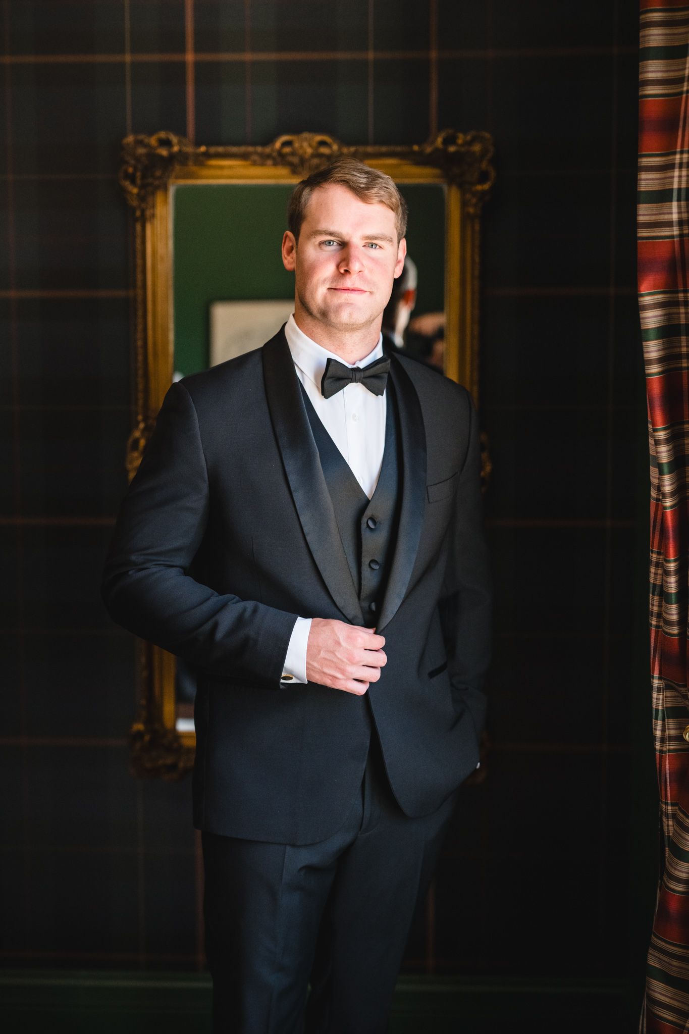 Dapper groom in black shawl lapel tuxedo with bow tie standing confidently before gold mirror and plaid wall.