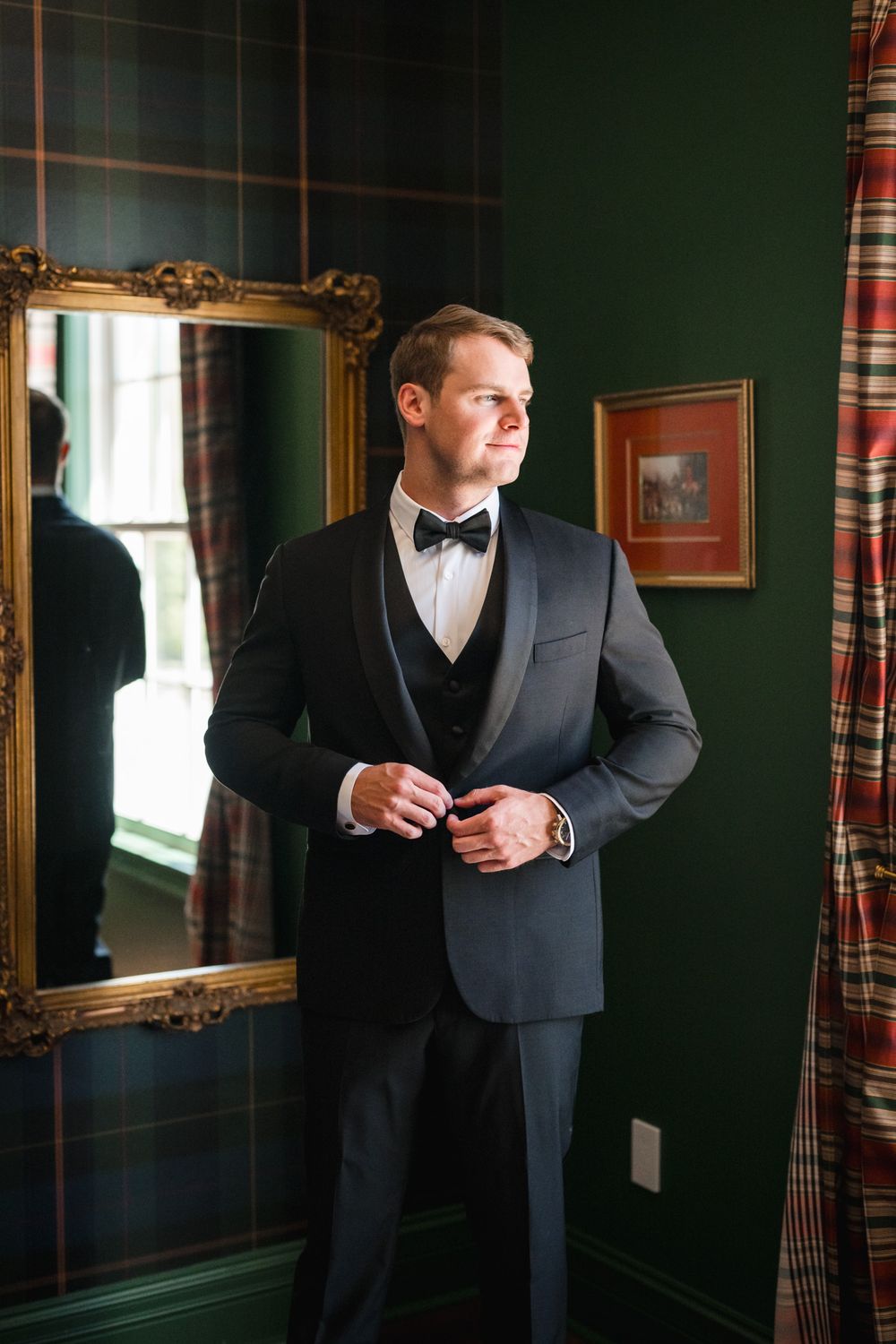 Groom in dark tuxedo with bow tie buttons jacket beside gold-framed mirror in green plaid room.