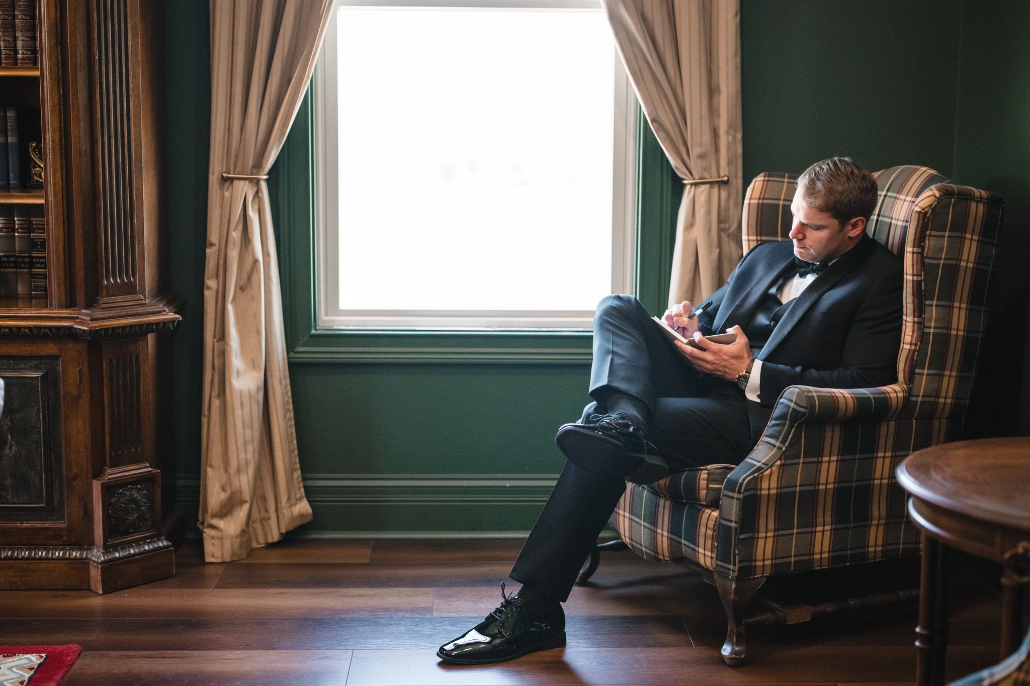 Groom in navy suit sitting in plaid armchair reading notes by window in green-walled room.