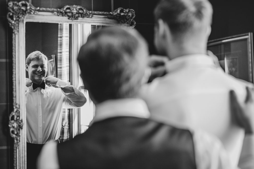 Groom adjusts bow tie in ornate mirror while groomsman assists, captured in classic black and white wedding photo.