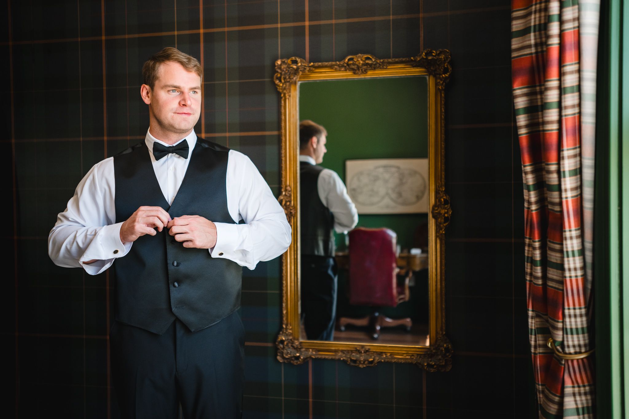 Groom in black tuxedo vest and bow tie buttoning jacket before gold mirror with plaid wall backdrop.