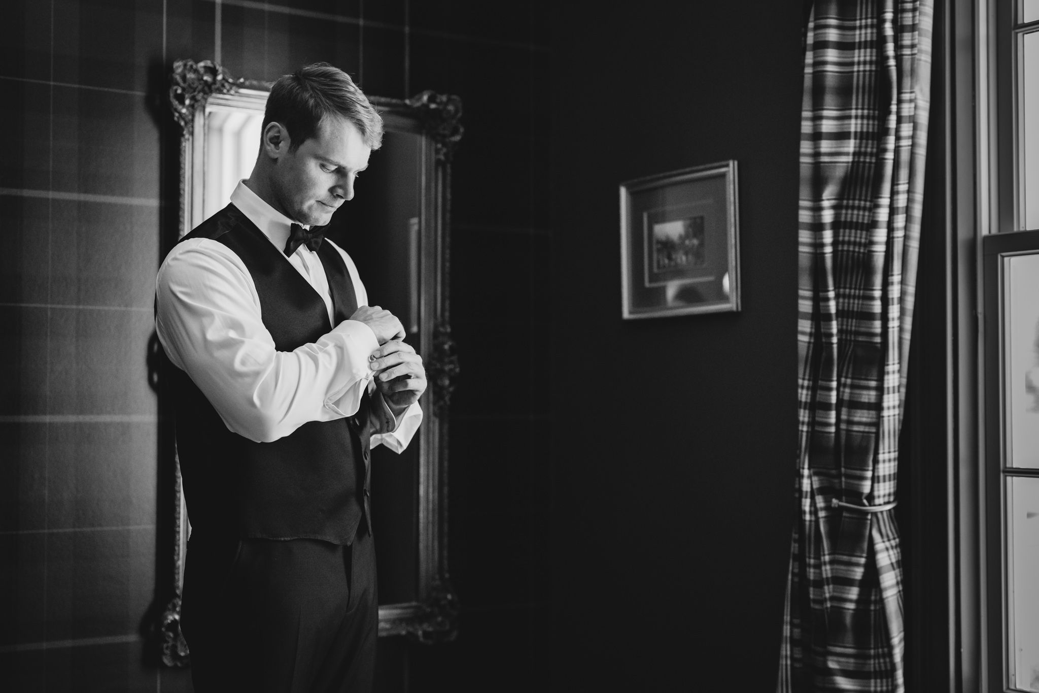 Groom in black vest and white shirt adjusting cufflinks in dimly lit room, black and white photo.