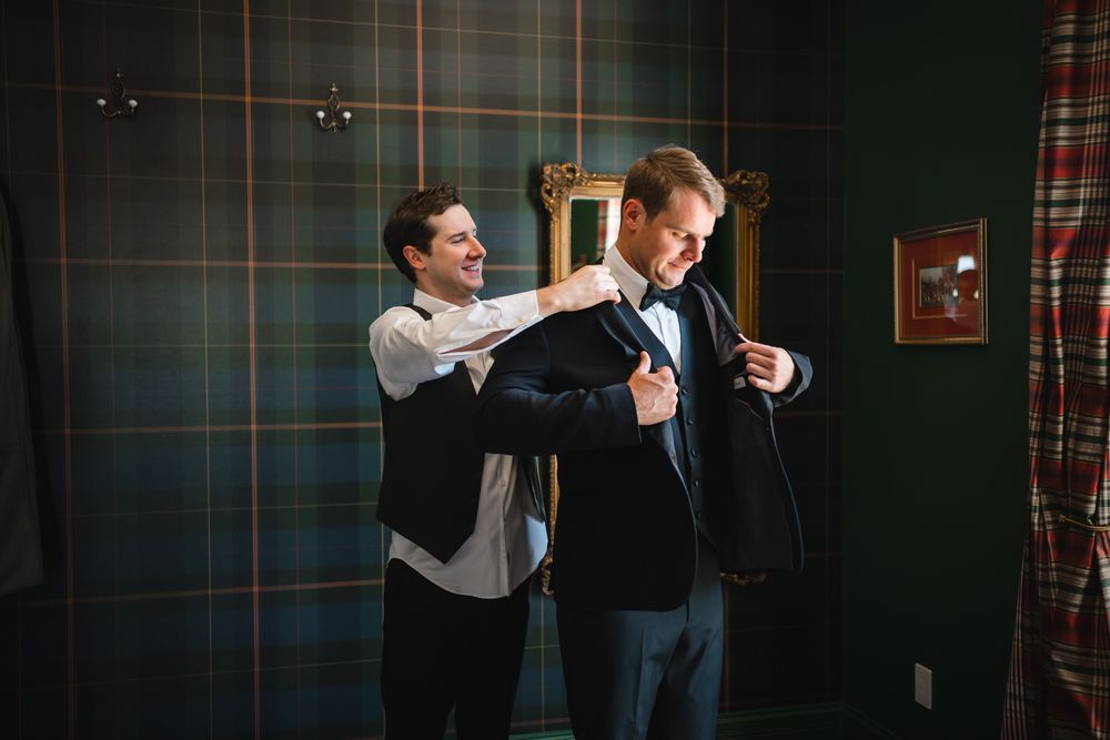 Groomsman helps groom adjust his black tuxedo jacket in a room with plaid tartan wallpaper.