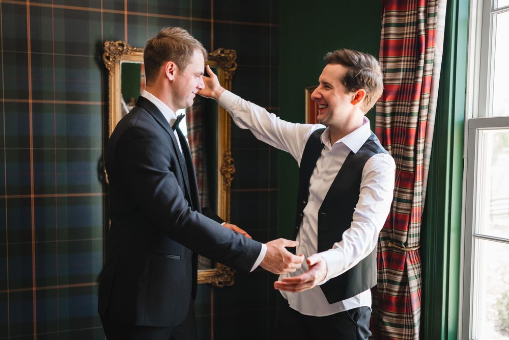 Two men laugh together as groomsman helps groom prepare in room with classic tartan plaid wallpaper.