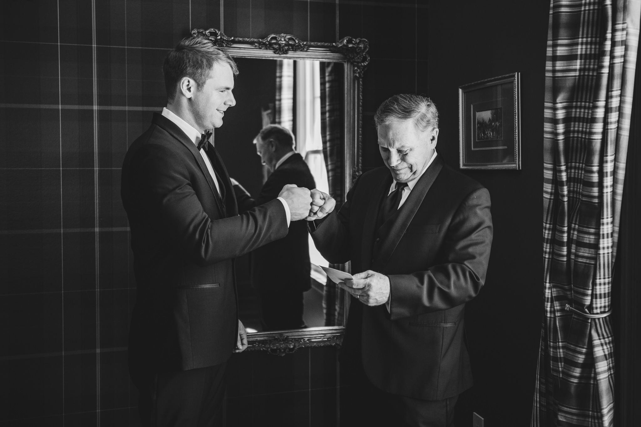 Black and white photo of groom and older man exchanging items in tartan room.