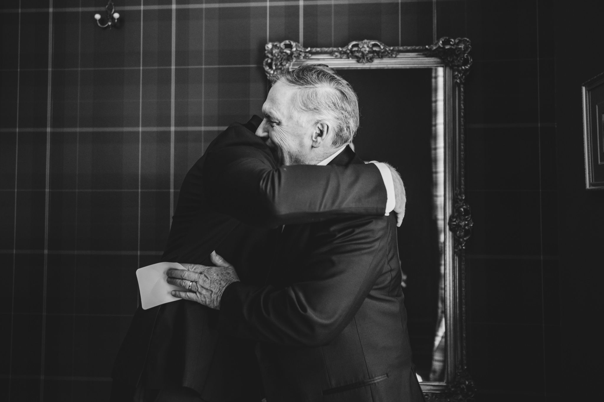 Two men share an emotional embrace in front of an ornate mirror in a tartan-walled room, black and white photo.