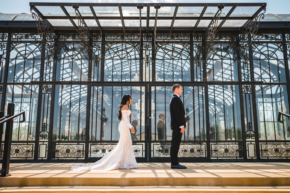 Bride and groom stand facing each other before ornate iron greenhouse gates in stunning wedding portrait.