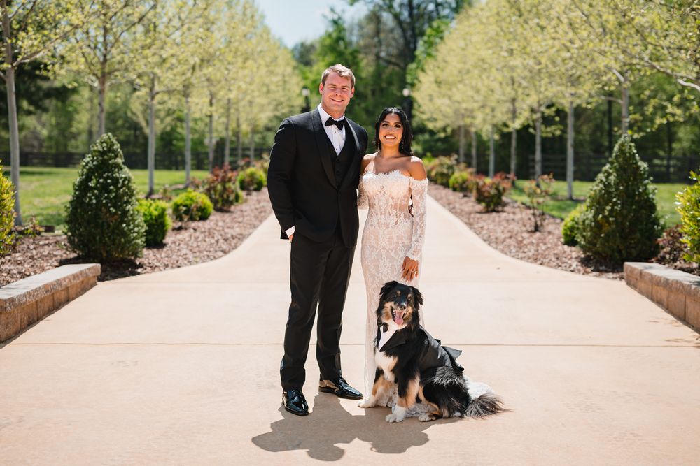 Wedding couple in formal attire pose with their dressed dog on a tree-lined venue pathway.