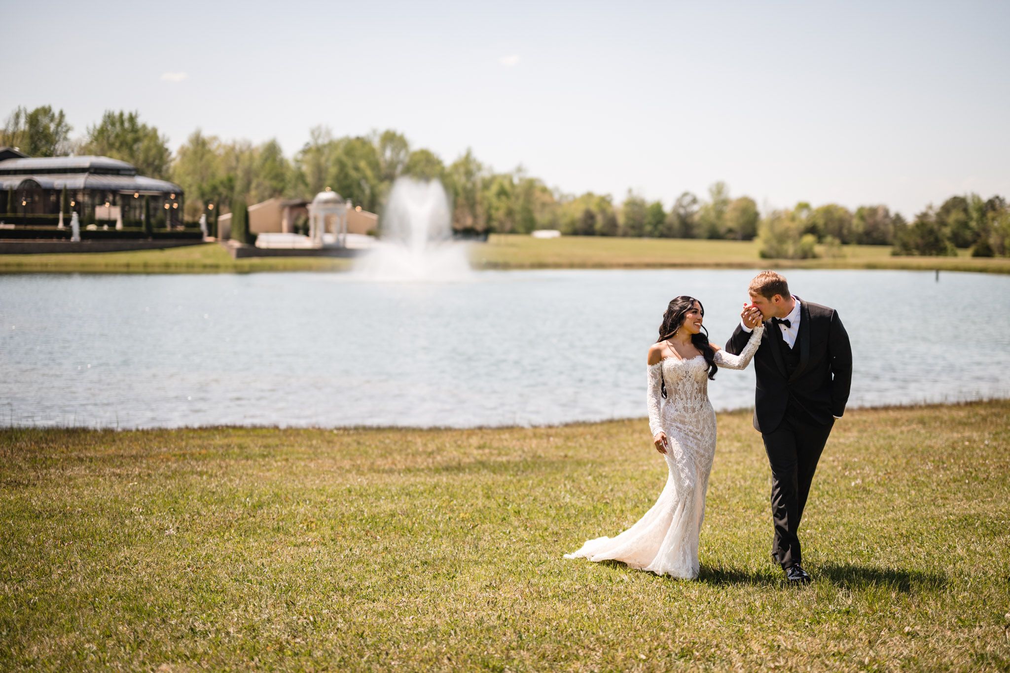 Bride and groom walk hand in hand by a serene lake with a fountain on their wedding day.