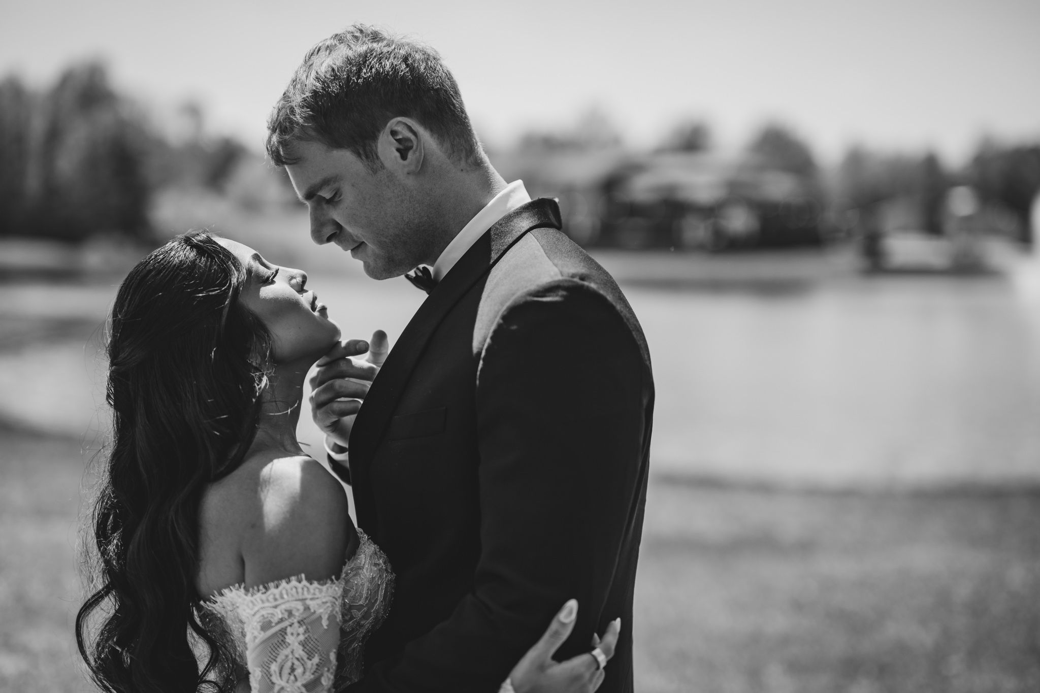 Romantic black and white photo of bride and groom embracing tenderly near a serene lakeside setting.
