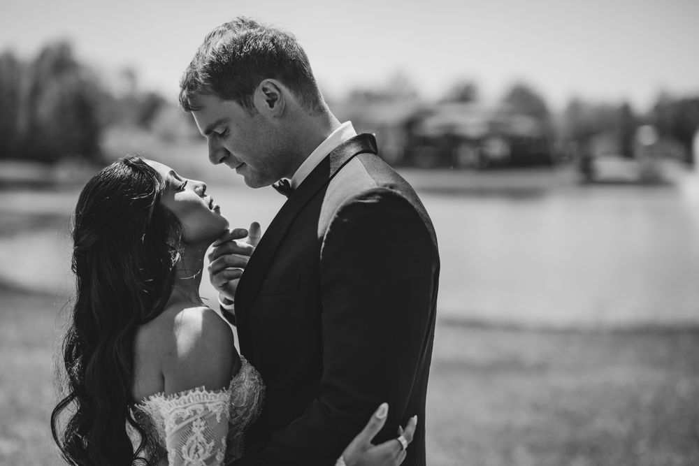 Romantic black and white photo of bride and groom embracing tenderly near a serene lakeside setting.