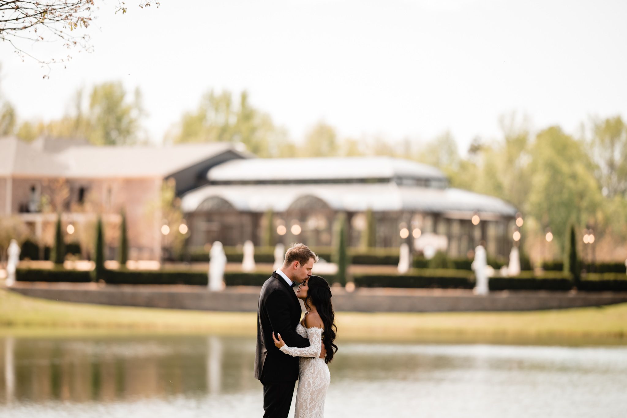 Newlyweds sharing a romantic kiss near a serene pond with a lit venue backdrop.