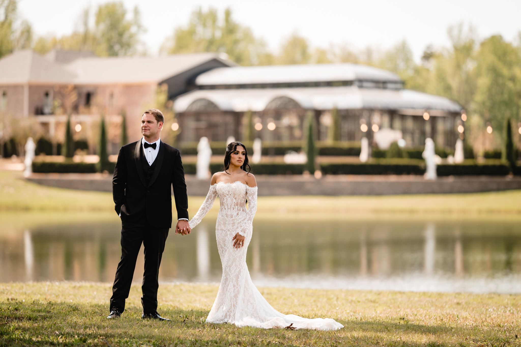 Bride and groom hold hands by a serene lake, bride in lace off-shoulder gown, groom in black tuxedo.