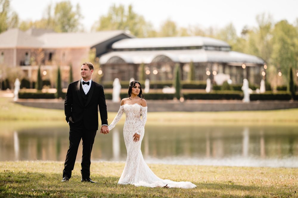 Bride and groom hold hands by a serene lake, bride in lace off-shoulder gown, groom in black tuxedo.