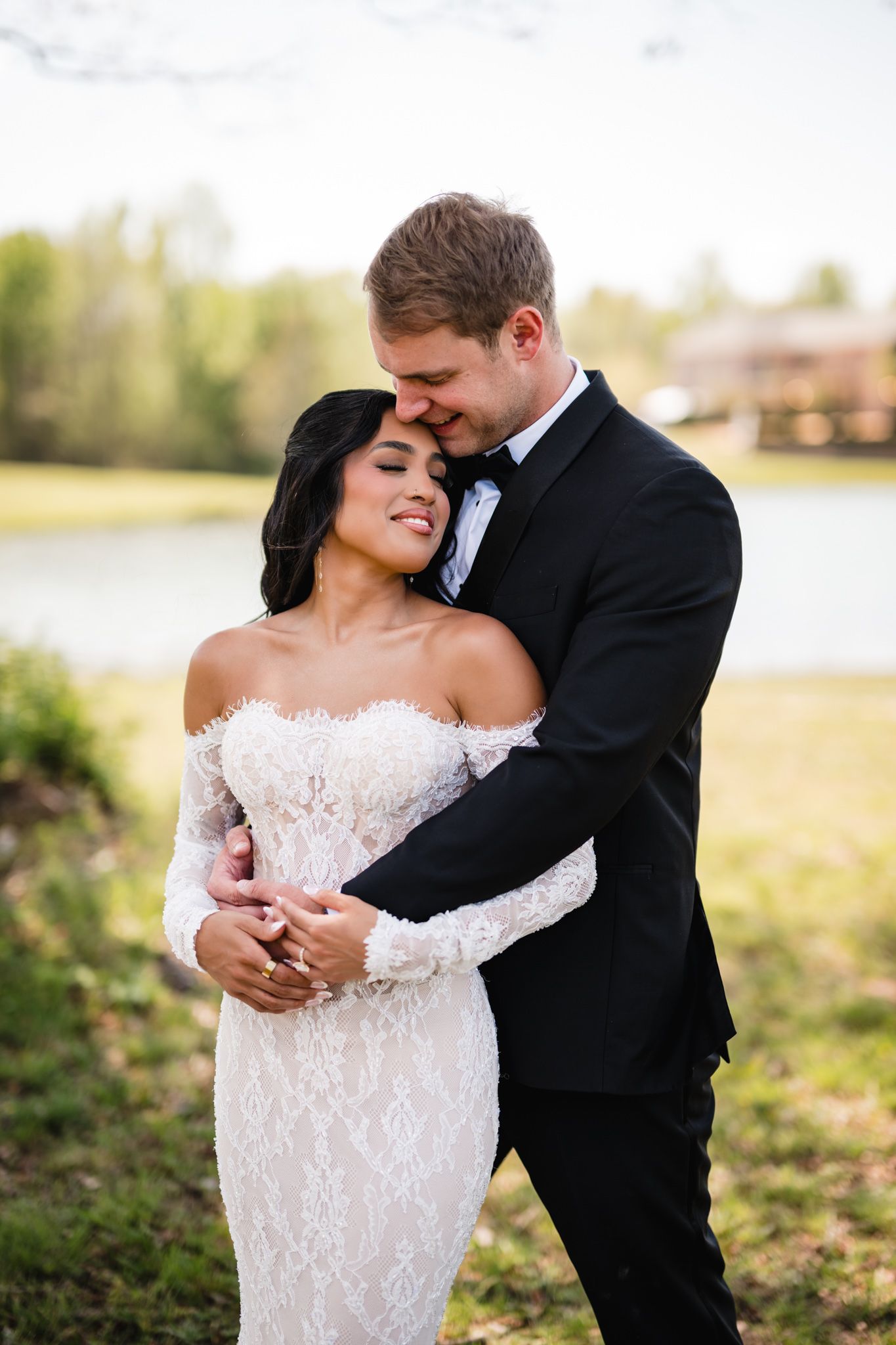 Groom in black tuxedo tenderly embraces smiling bride in lace off-shoulder gown outdoors near serene pond.