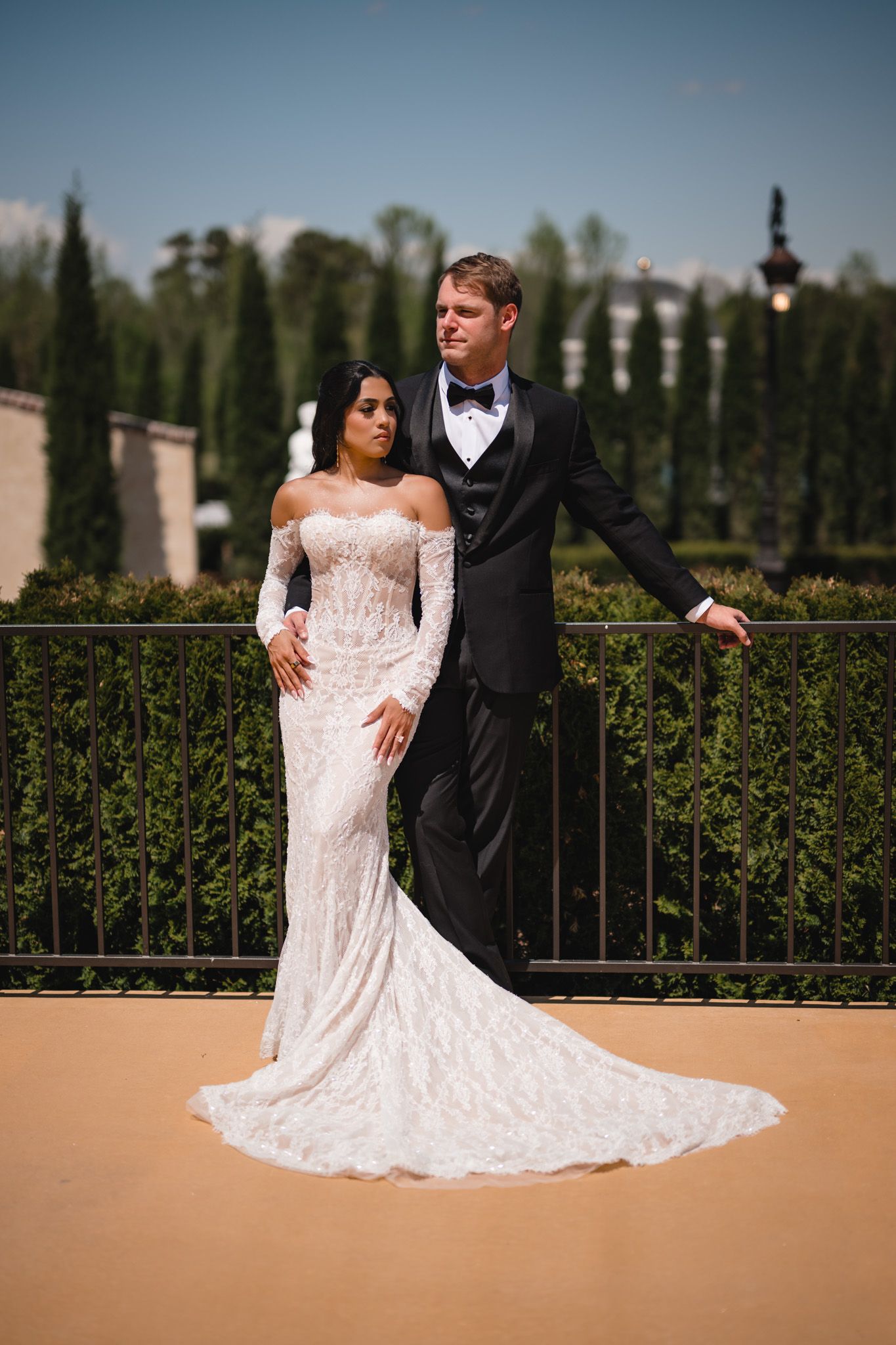 Elegant bride in off-shoulder lace gown stands with tuxedo-clad groom on a terrace with cypress trees.