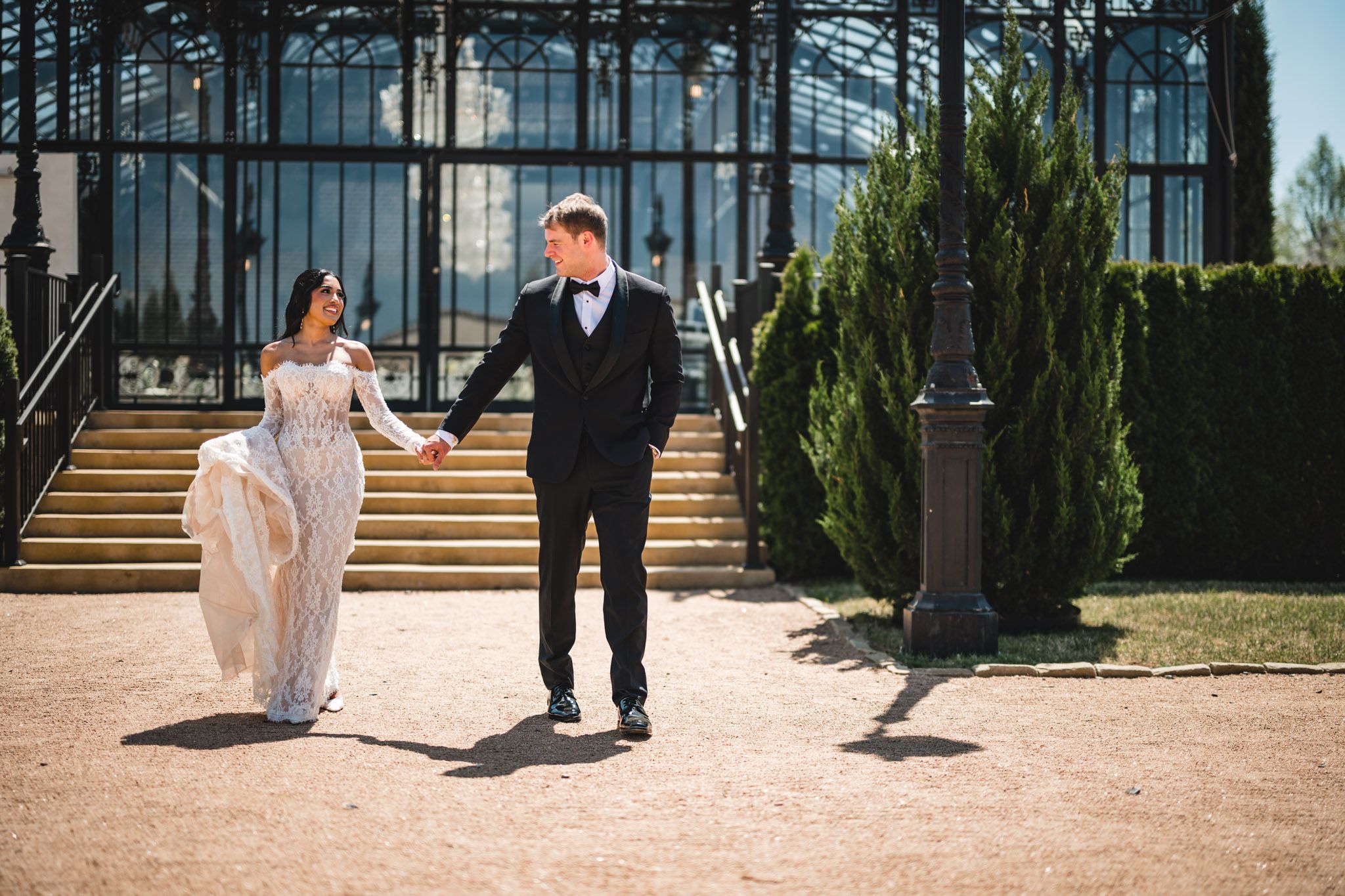 Bride and groom walk hand in hand before a grand glass greenhouse on their wedding day.