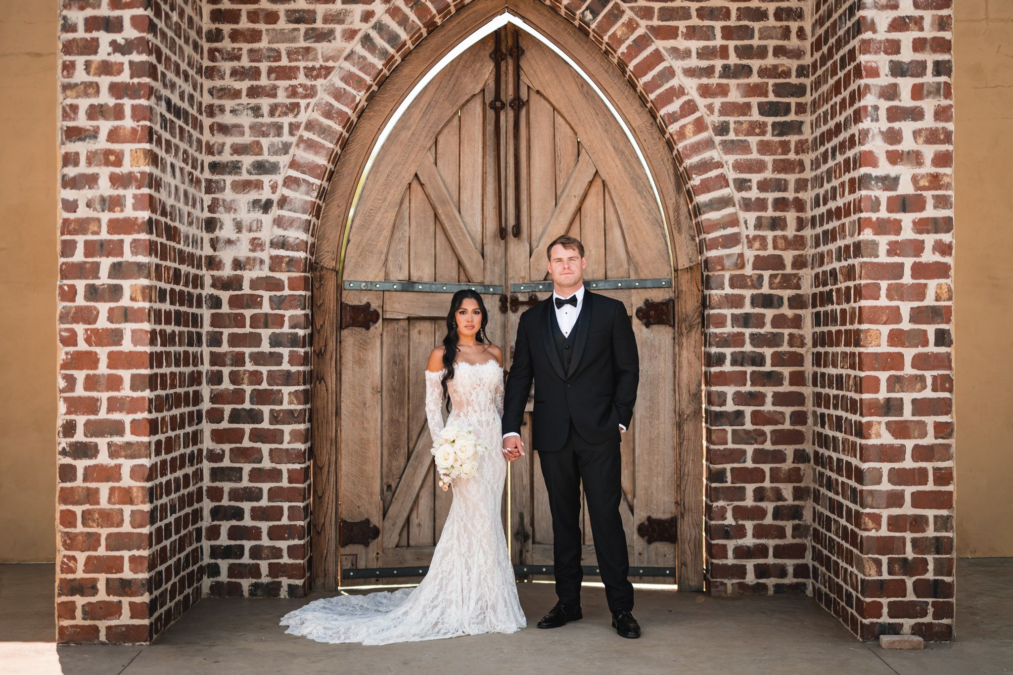 Bride and groom pose together under a gothic brick archway with wooden doors at their wedding venue.