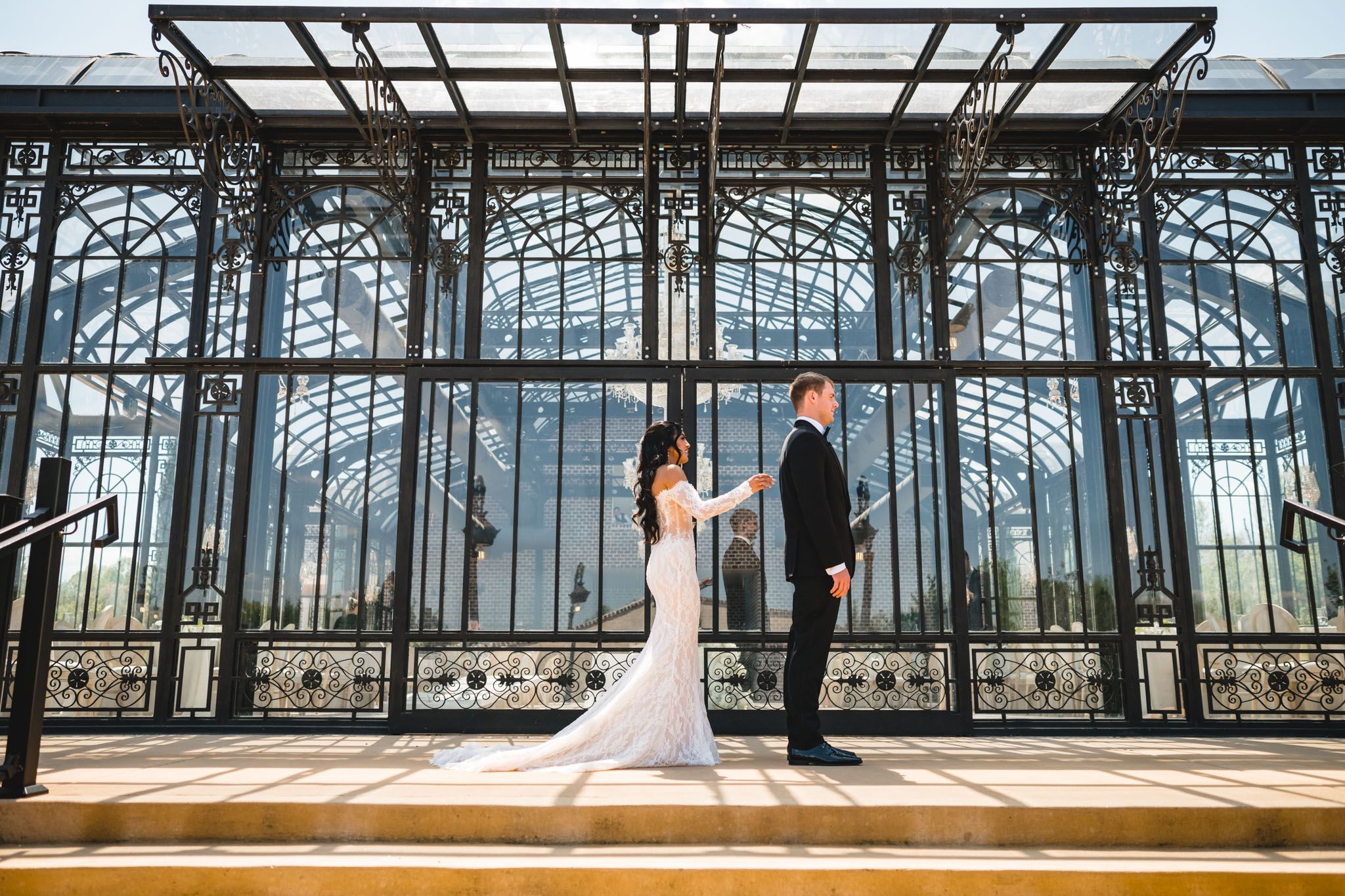 Bride and groom hold hands in front of an ornate iron and glass greenhouse structure on their wedding day.
