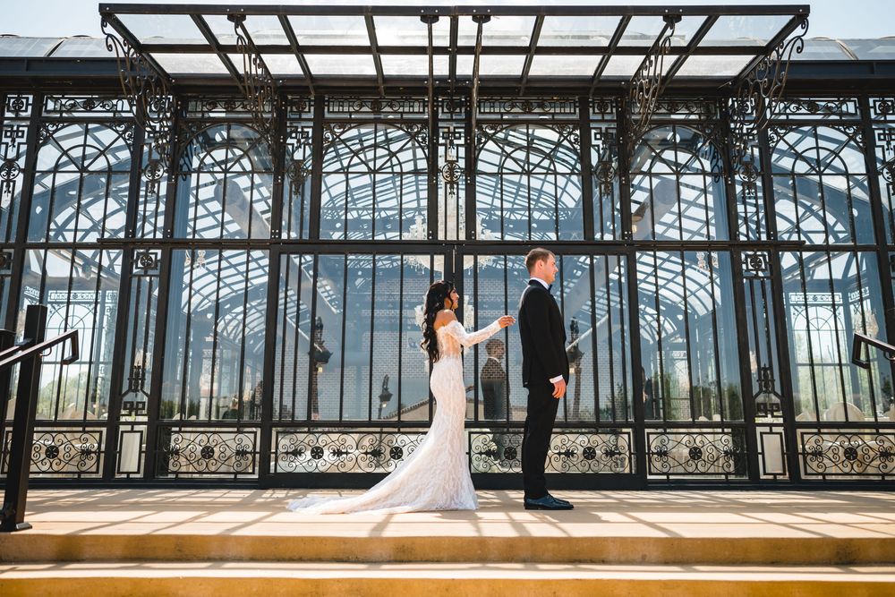 Bride and groom hold hands in front of an ornate iron and glass greenhouse structure on their wedding day.